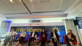 Six people are sitting on chairs. Behind them is a banner with the text "11th annual european Cybersecurity conference"
