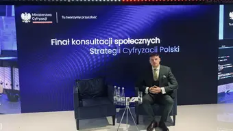 A person sitting on stage. Behind him graphic by Polish Ministry of Digital Affairs